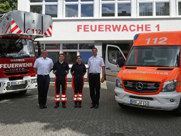 Vier Rettungsdienstmitarbeiter vor der Rettungswache. Links steht die Drehleiter und rechts der Rettungwagen 
