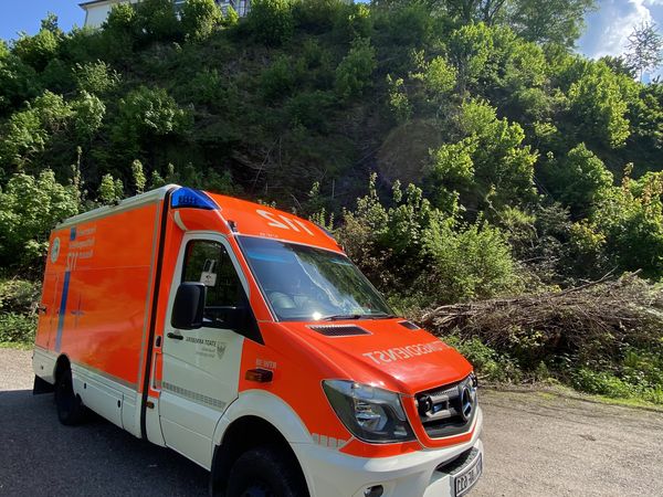 Ein Rettungswagen vor einem Gebüsch an einem sonnigen Tag. Im Hintergrund eine Villa mit Rundturm erkennbar