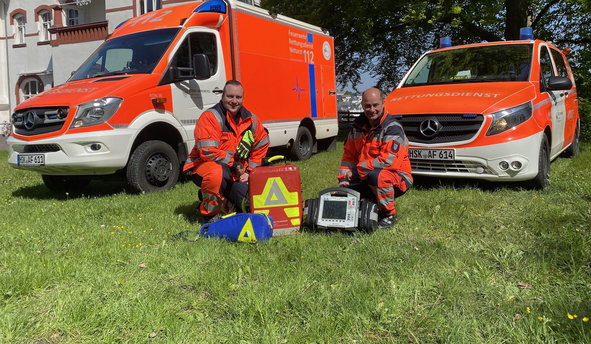Zwei Rettungsdienstmitarbeiter kniend im Gras mit Geräten des Rettungsdienstes. Im Hintergrund ein RTW und NEF